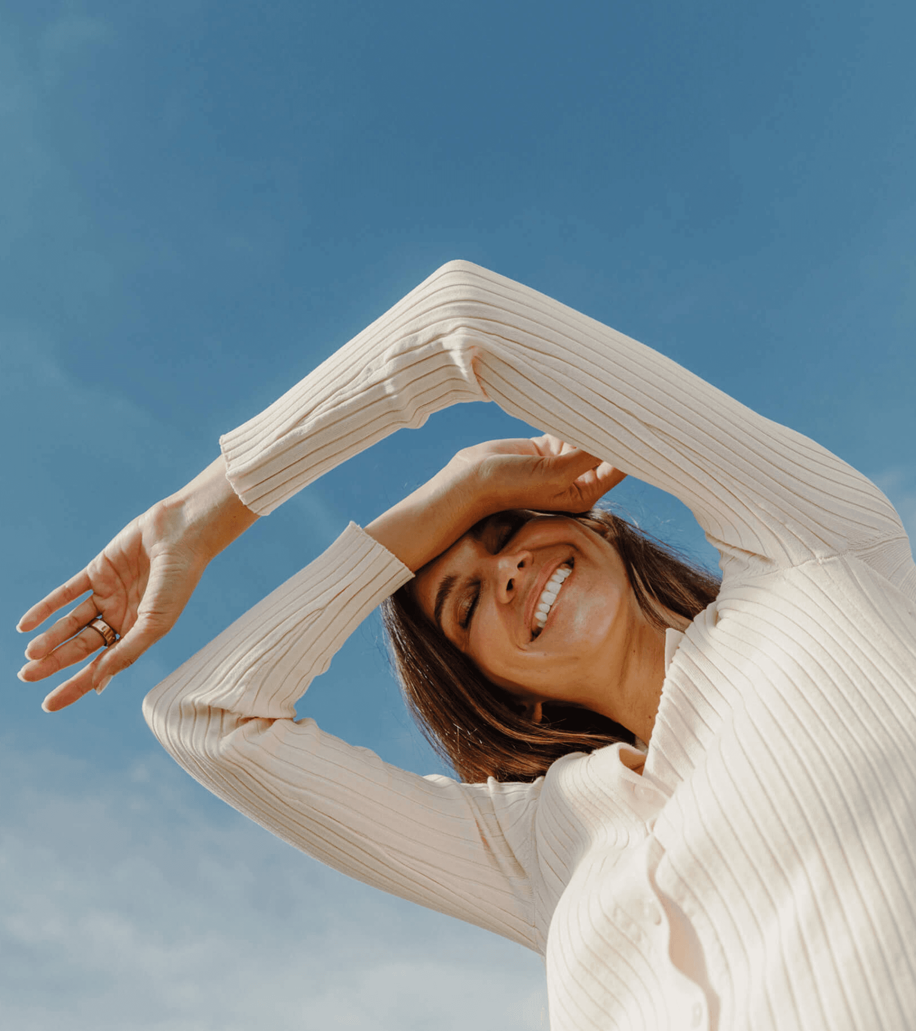 A woman in an ivory knit top stretching in the sunshine beneath a bright blue sky